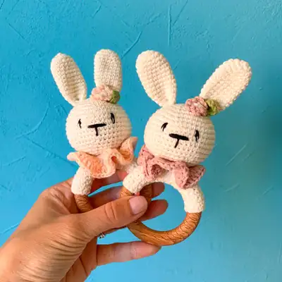 Two completed bunny baby rattle amigurumi pieces in cream yarn with pink collars held by a hand, showing front view of heads and wooden teether rings