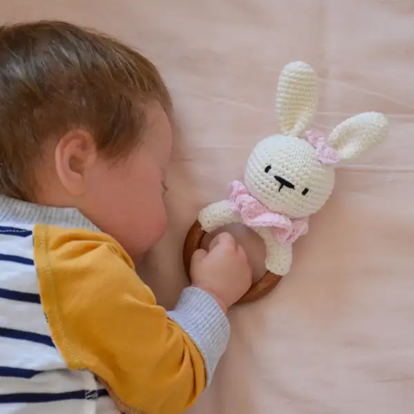 Sleeping baby reaching for a bunny baby rattle amigurumi lying beside them, showing scale and ring size